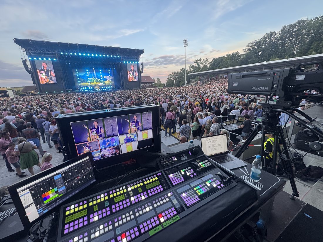 Regiepult mit Multiviewer vor einer Open-Air-Bühne, im Hintergrund Publikum und Bühne bei Abendlicht