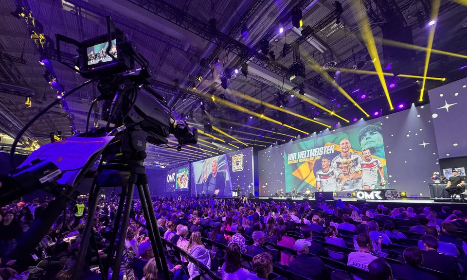 Großkonferenz in einer Messehalle: Kamera auf Stativ im Vordergrund, Bühne mit LED-Wand und Publikum